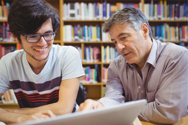 A young man and an older man chat in front of a bookcase with a laptop.